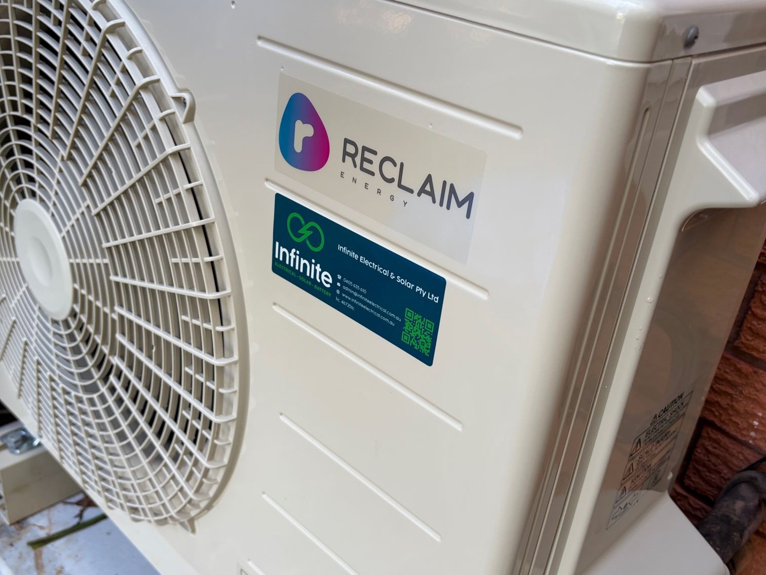 Reclaim Energy heat pump outdoor unit installed against a brick wall, with an Infinite Electrical & Solar sticker