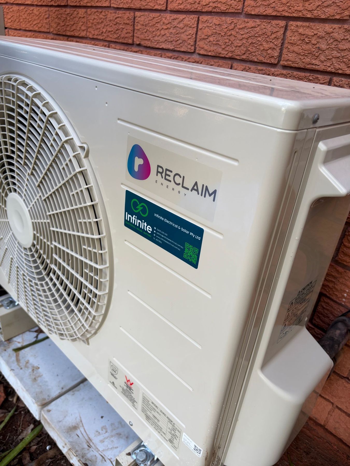 Reclaim Energy heat pump outdoor unit installed against a brick wall, showing the entire unit with an Infinite Electrical & Solar sticker